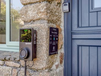 An electric vehicle charging point mounted on a stone wall next to a blue door at The Cubby Hole in Sennen Cove