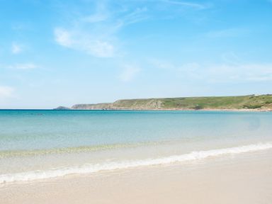 A sandy beach with clear water and a grassy headland in the background at The Cubby Hole in Sennen Cove