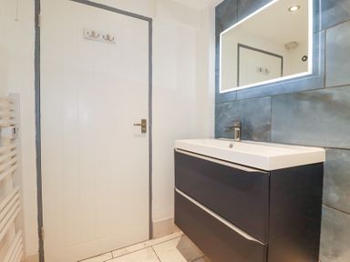 A bathroom with a white door a dark vanity with a white sink and a lit mirror above at The Cubby Hole in Sennen Cove