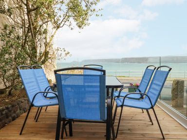 An outdoor deck with a glass table and six blue chairs overlooking the sea at The Cubby Hole in Sennen Cove