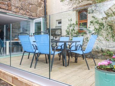 An outdoor wooden deck with blue chairs around a table enclosed by glass panels with a stone wall in the background at The Cubby Hole in Sennen Cove