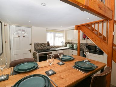 A dining area with a wooden table set with teal plates and wine glasses next to a kitchen with white cabinets and a wooden staircase at Harvest Moon Cottage in Much Cowarne