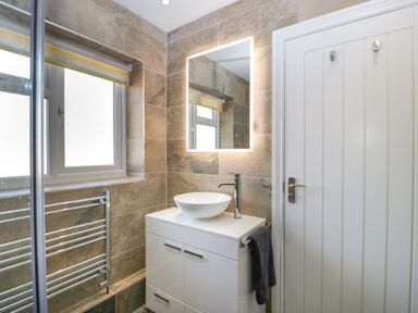 A bathroom with a white vanity cabinet with a vessel sink a lit mirror a window with a yellow blind and a towel rail at Harvest Moon Cottage in Much Cowarne