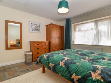 A bedroom with a wooden wardrobe, chest of drawers, mirror, window with curtains, and a bed with green patterned bedding at Harvest Moon Cottage in Much Cowarne