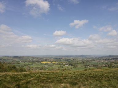 A grassy hill overlooking a wide landscape with fields trees and scattered houses under a partly cloudy sky at Harvest Moon Cottage in Much Cowarne
