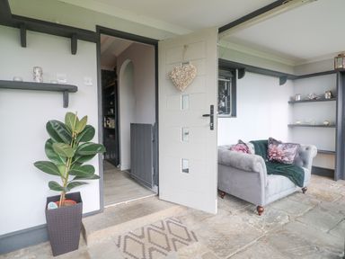 An open door leading into a room with a grey armchair with floral pillows a potted plant near the door shelves on the walls and a stone floor at The Lodge at Wildersley Farm in Belper