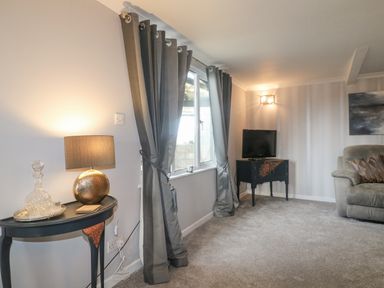 A living room with a round table with lamp and glassware near window with curtains and a television on a small table at The Lodge at Wildersley Farm in Belper