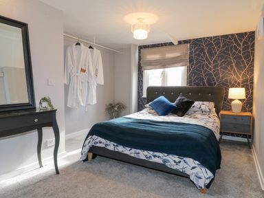 A bedroom with a bed blue and white bedding a bedside table with a lamp robes hanging a mirror on a black dressing table at The Lodge at Wildersley Farm in Belper