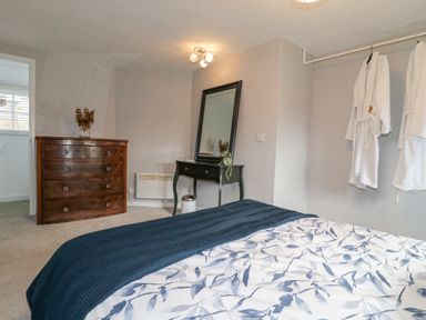 A bedroom with a bed covered in a blue and white patterned blanket a wooden chest of drawers a black dressing table with a mirror and two white robes hanging on a rail at The Lodge at Wildersley Farm in Belper