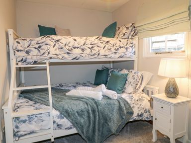 A bedroom with a white bunk bed, floral bedding, pillows, a green blanket, white rolled towels, a nightstand with a lamp, and a window at The Lodge at Wildersley Farm in Belper