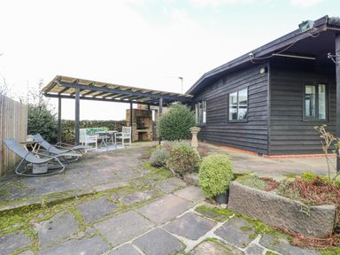 An outdoor patio area with a wooden pergola dining set lounge chairs and a barbecue next to a black wooden house at The Lodge at Wildersley Farm in Belper