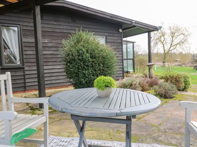An outdoor patio with a round wooden table and two white chairs in front of a dark wooden house with bushes at The Lodge at Wildersley Farm in Belper