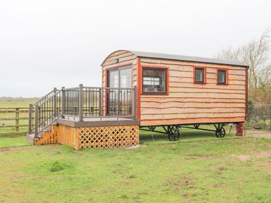 A wooden shepherd hut on wheels with a small deck and railing in a grassy field at Tal Sarn Shepherd Hut in Dwyran near Niwbwrch Newborough