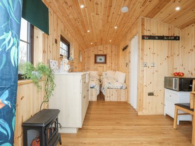 A small wooden room with two single beds a kitchenette with a sink a microwave on a small cabinet and a black heater at Tal Sarn Shepherd Hut in Dwyran near Niwbwrch Newborough