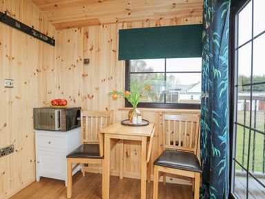 A small wooden dining area with two chairs a table a microwave on a cabinet and a window with curtains at Tal Sarn Shepherd Hut in Dwyran near Niwbwrch Newborough
