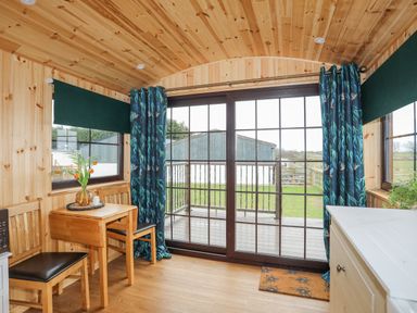 A wooden room with a small table and two chairs near large glass sliding doors with blue patterned curtains at Tal Sarn Shepherd Hut in Dwyran near Niwbwrch Newborough