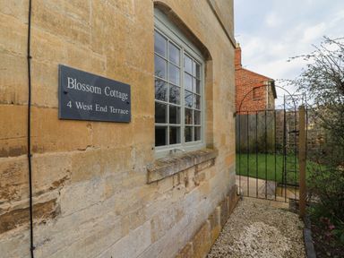 The exterior wall of a stone building with a window a sign saying Blossom Cottage and a metal garden gate at Blossom Cottage in Chipping Campden