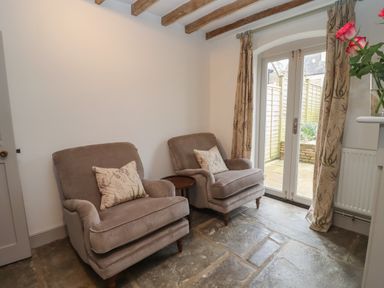 A room with two armchairs, a small round wooden table, floral curtains, and glass doors leading outside at Blossom Cottage in Chipping Campden