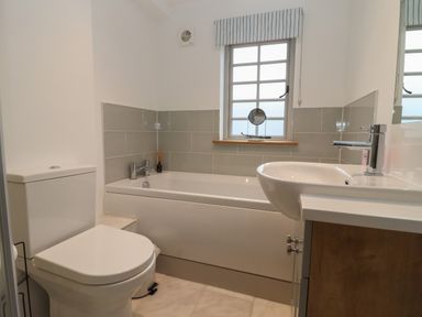 A bathroom with a toilet a bathtub with tiles around and a sink below two windows at Blossom Cottage in Chipping Campden