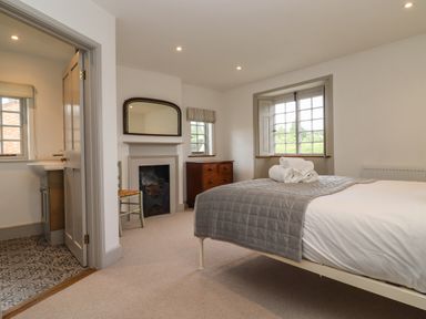 A bedroom with a bed covered by a gray quilt and rolled towels on top a fireplace with a mirror above a wooden chair a wooden dresser and a window at Blossom Cottage in Chipping Campden