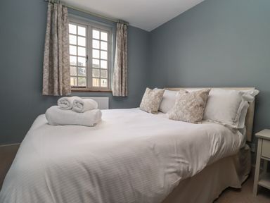 A bedroom with a white bed with rolled towels on it beige patterned cushions and a window with beige curtains at Blossom Cottage in Chipping Campden