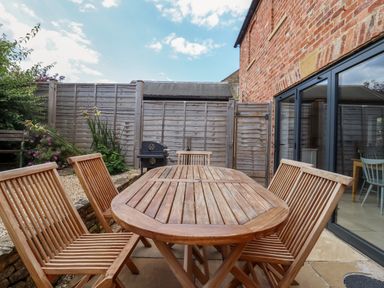 An outdoor patio area with a wooden table and chairs a barbecue grill and a brick wall with sliding glass doors at Blossom Cottage in Chipping Campden