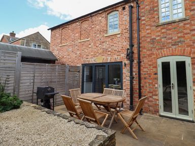 An outdoor patio area with a wooden table and chairs a barbecue grill and brick house walls at Blossom Cottage in Chipping Campden