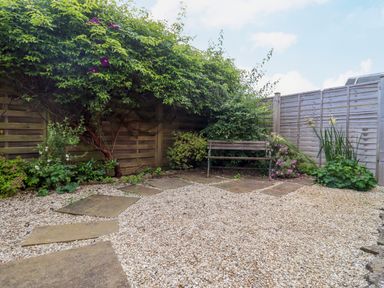 A garden area with gravel ground and stepping stones leading to a wooden bench surrounded by plants and a wooden fence at Blossom Cottage in Chipping Campden