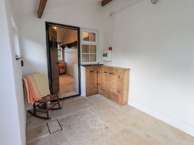A small room with a wooden rocking chair draped with a striped blanket a wooden corner cabinet and an open door leading to another room at Green Yew in Winster near Bowness-On-Windermere