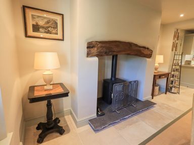 A room with a wood stove fireplace with a rustic wooden mantel a small table with a lamp and a framed picture on the wall at Green Yew in Winster near Bowness-On-Windermere