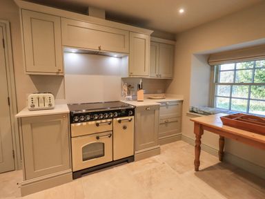 A kitchen with beige cabinets a cream oven a white toaster a wooden table and a window with a cushion on the sill at Green Yew Winster near Bowness-On-Windermere