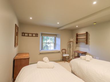 A bedroom with two beds a wooden chest a window with a cushion a chair a coat stand and a wooden shelf at Green Yew in Winster near Bowness-On-Windermere