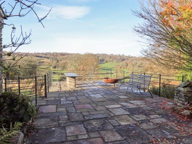 An outdoor patio with stone paving a metal bench a fire pit and scenic countryside view at Green Yew in Winster near Bowness-On-Windermere