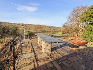 A stone patio with a long rectangular stone table and a metal fire pit surrounded by metal railings at Green Yew in Winster near Bowness-On-Windermere