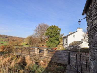 An outdoor stone patio with a metal railing a bench and a table near a white house in a rural area at Green Yew in Winster near Bowness-On-Windermere