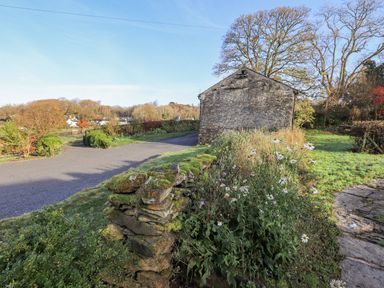 A stone building next to a road with a moss-covered stone wall and wildflowers at Green Yew in Winster near Bowness-On-Windermere