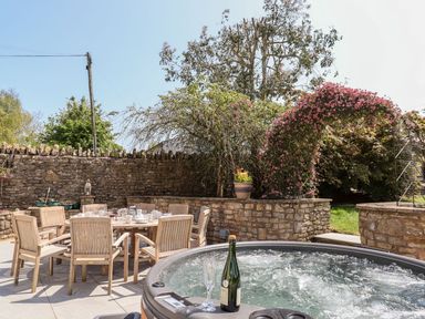 An outdoor patio with a wooden dining table and chairs set with dishes and a hot tub with a bottle and glass against a stone wall with plants at Nempnett Farmhouse in Nempnett Thrubwell near Blagdon