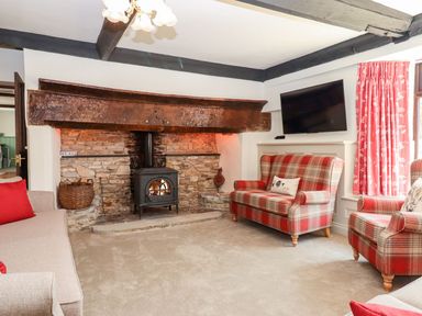 A living room with a stone fireplace and wood stove with plaid armchairs and a wall-mounted television at Nempnett Farmhouse in Nempnett Thrubwell near Blagdon