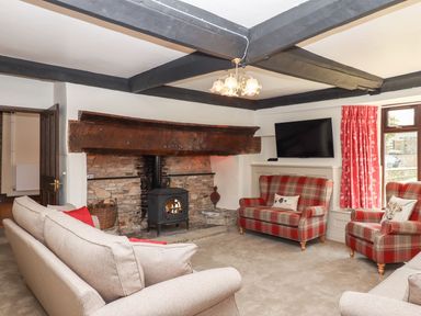 A living room with a stone fireplace and wood-burning stove two beige sofas and two red plaid armchairs with cushions a wall-mounted television and red curtains at Nempnett Farmhouse in Nempnett Thrubwell near Blagdon