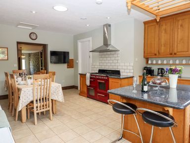 A kitchen with a dining table and wooden chairs a red stove and a kitchen island with stools a bottle and flowers at Nempnett Farmhouse in Nempnett Thrubwell near Blagdon