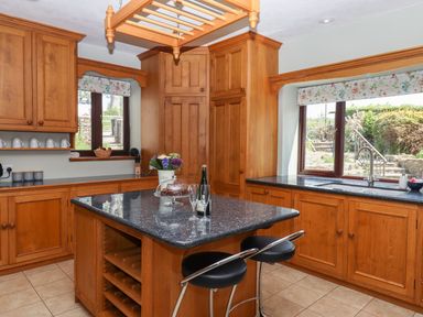 A kitchen with wooden cabinets a black countertop an island with wine bottles and glasses and windows overlooking a garden at Nempnett Farmhouse in Nempnett Thrubwell near Blagdon