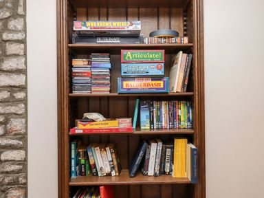 A wooden bookshelf with board games puzzles cds dvds and books next to a stone wall at Nempnett Farmhouse in Nempnett Thrubwell near Blagdon