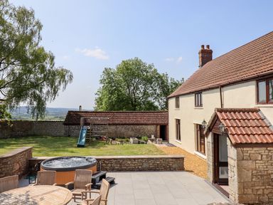 A backyard with a round hot tub a wooden table with chairs a slide and a two story house with a tiled roof at Nempnett Farmhouse in Nempnett Thrubwell near Blagdon