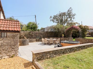 An outdoor patio area with a dining table and chairs a round hot tub stone walls and a pathway at Nempnett Farmhouse in Nempnett Thrubwell near Blagdon