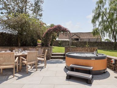 An outdoor patio area with wooden dining table and chairs and a hot tub with steps surrounded by a stone wall at Nempnett Farmhouse in Nempnett Thrubwell near Blagdon