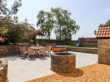 An outdoor patio area with a round wooden table and chairs a stone well a hot tub a green slide and trees at Nempnett Farmhouse in Nempnett Thrubwell near Blagdon