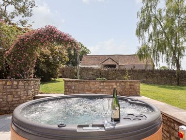 An outdoor hot tub with a bottle of wine and two glasses next to a stone wall and garden at Nempnett Farmhouse in Nempnett Thrubwell near Blagdon