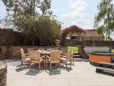 An outdoor patio with a wooden dining table and chairs set with dishes and a hot tub on tiled flooring at Nempnett Farmhouse in Nempnett Thrubwell near Blagdon