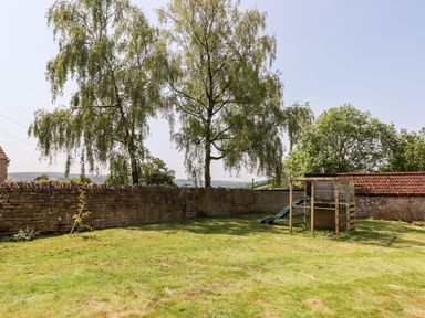 A garden with green grass, two large trees, a stone wall, and a wooden play structure with a slide at Nempnett Farmhouse in Nempnett Thrubwell near Blagdon
