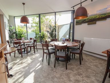 A dining area with round wooden tables and chairs in front of large windows and plants at Lodge 3 in St Asaph